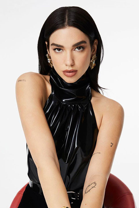 Dua Lipa in full black and she is sitting on a chair